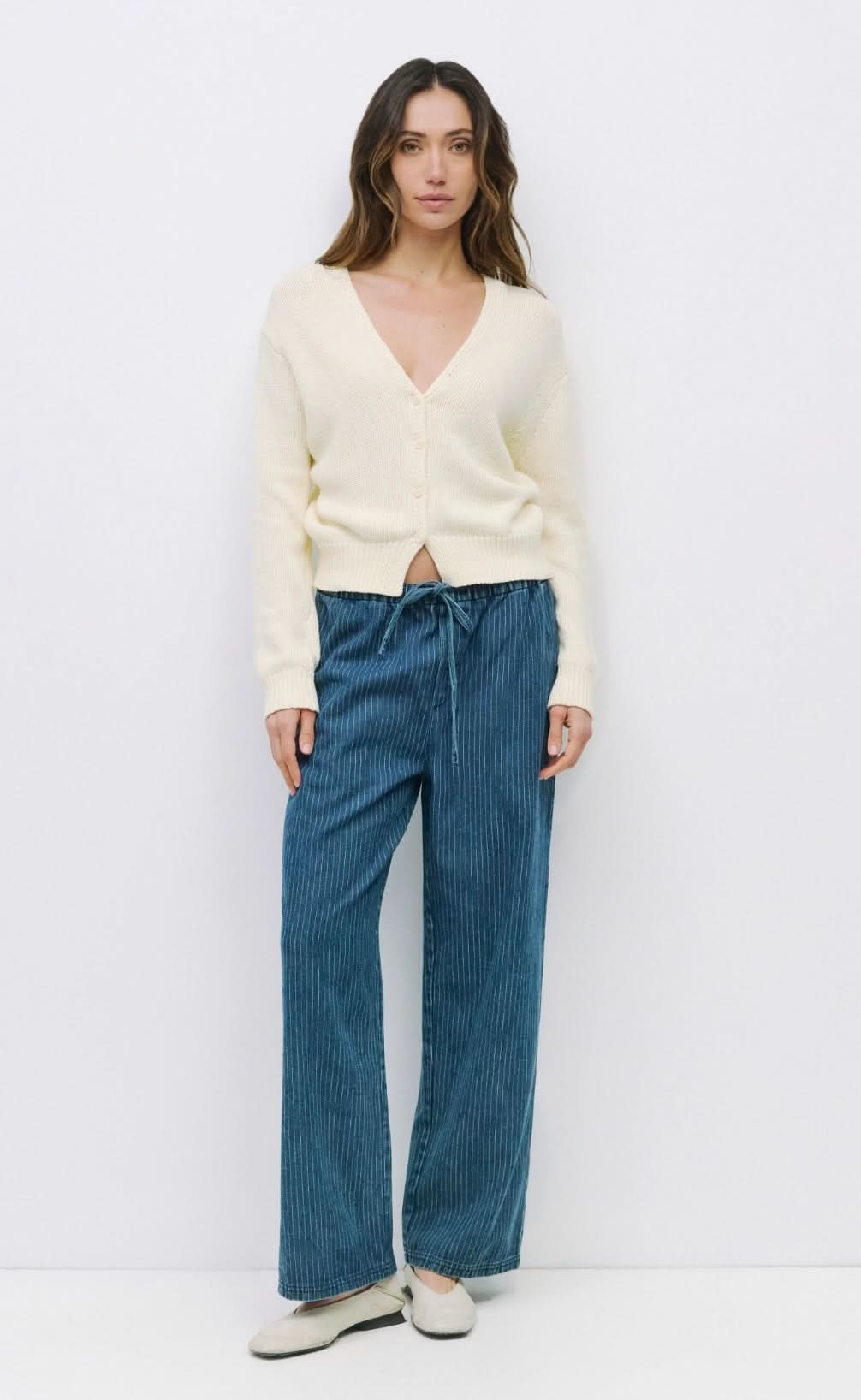 Woman wearing off white knit long sleeve sweater deep v neck with front button closure paired with pinstriped blue drawstring jeans