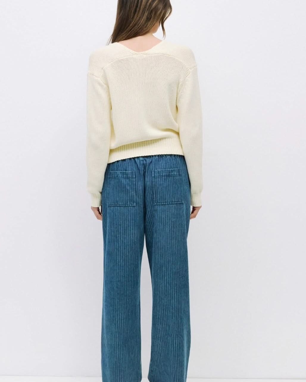 Woman facing backward wearing off white knit long sleeve sweater paired with pinstriped blue drawstring jeans