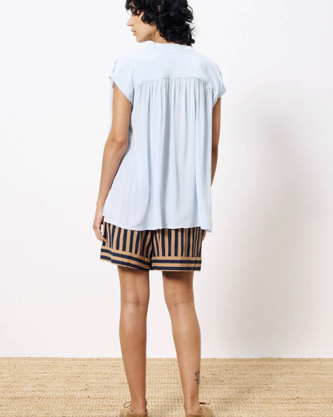 Woman turned backward wearing lightweight light blue blouse paired with tan and black dress shorts