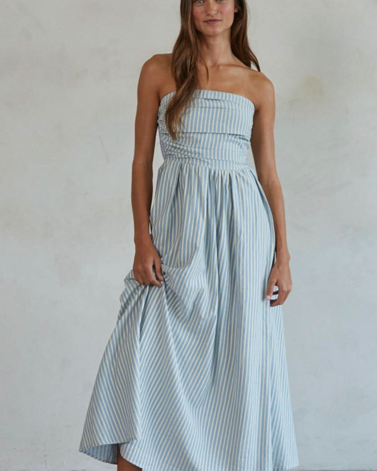 Woman wearing white and green/blue vertically striped, sleeveless maxi dress standing against a blank wall