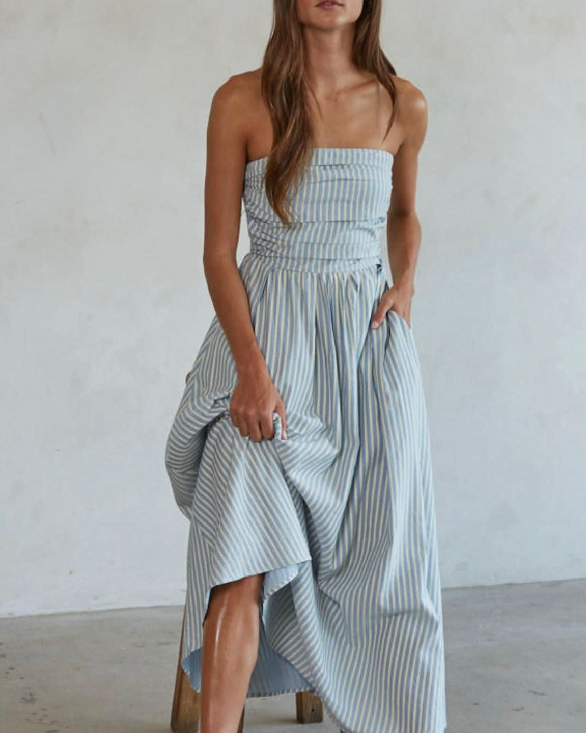 Woman wearing white and green/blue vertically striped, sleeveless maxi dress standing against a blank wall