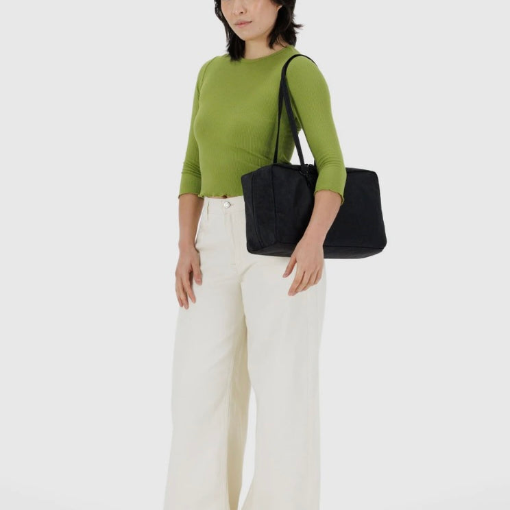 Woman wearing a green top and white pants holding a black BAGGU bag on a white background