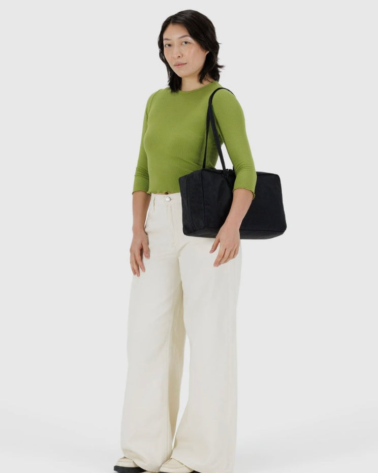 Woman wearing a green top and white pants holding a black BAGGU bag on a white background
