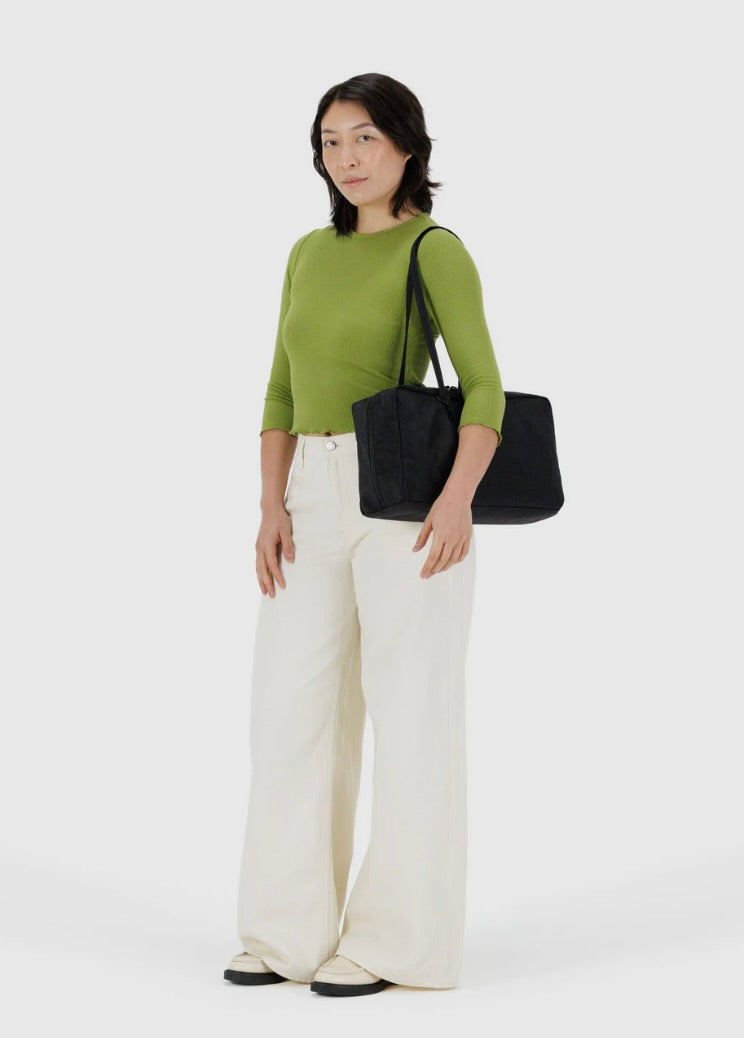 Woman wearing a green top and white pants holding a black BAGGU bag on a white background