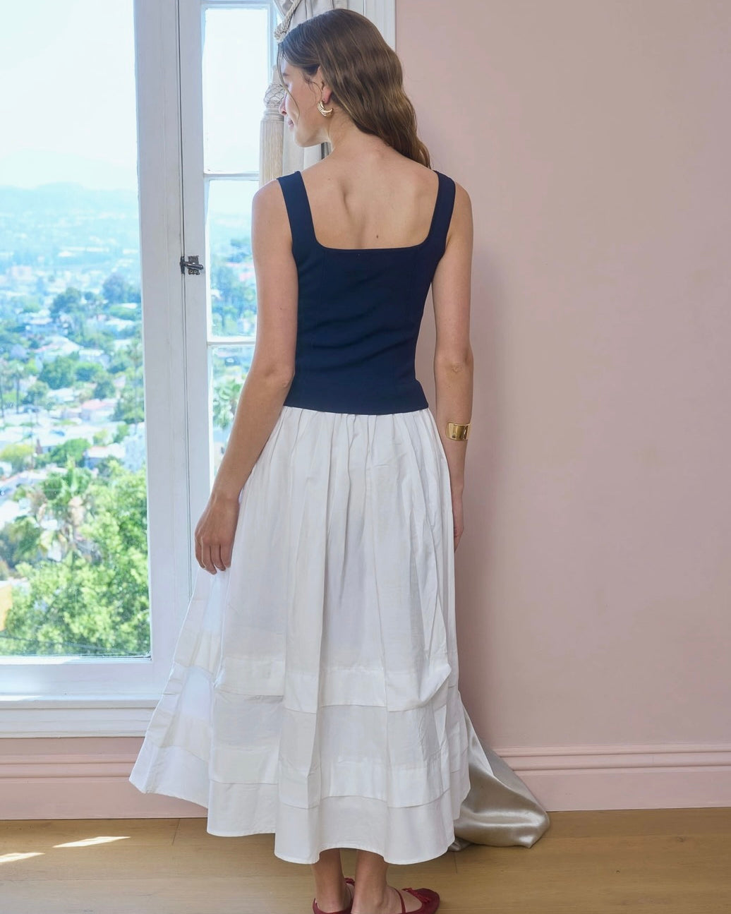 Woman wearing a navy top and white skirt standing by a window with a view of trees.