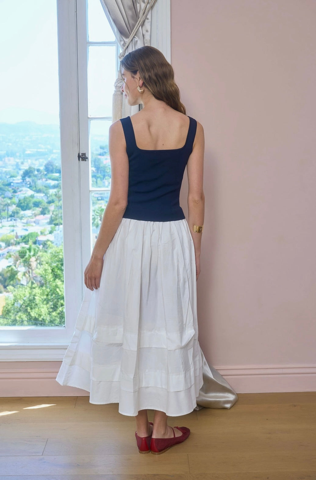 Woman wearing a navy top and white skirt standing by a window with a view of trees.