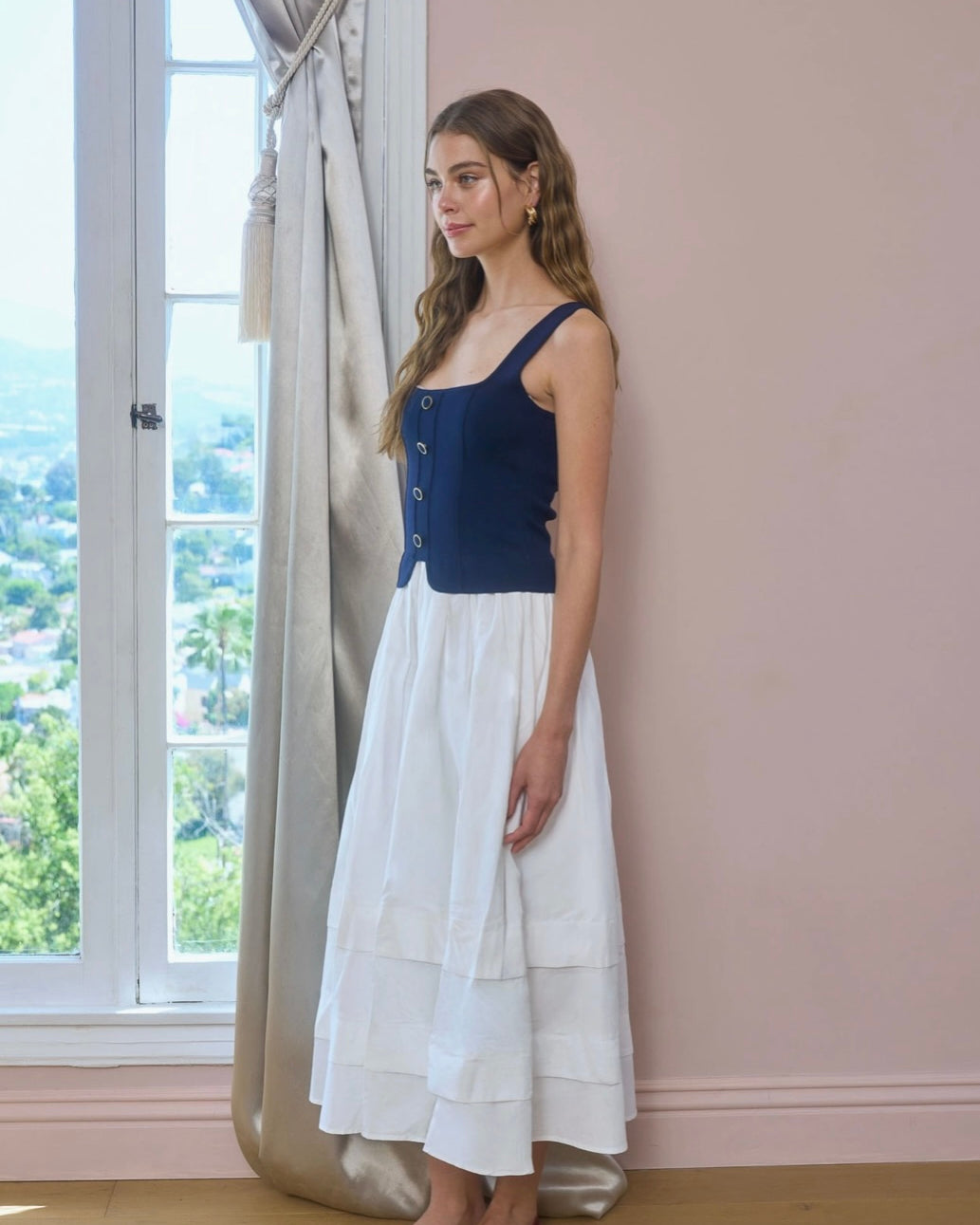 Woman wearing a blue top and white skirt standing in a room with large windows.