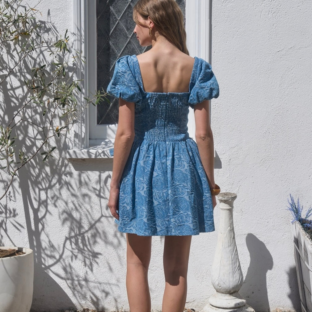 Woman wearing a blue dress standing outdoors against a white wall.