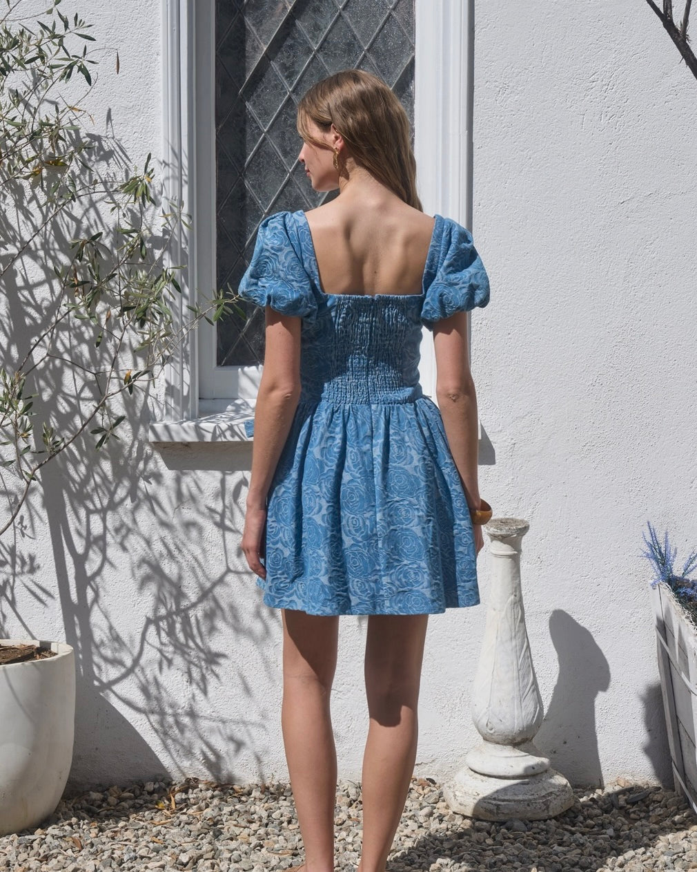 Woman wearing a blue dress standing outdoors against a white wall.
