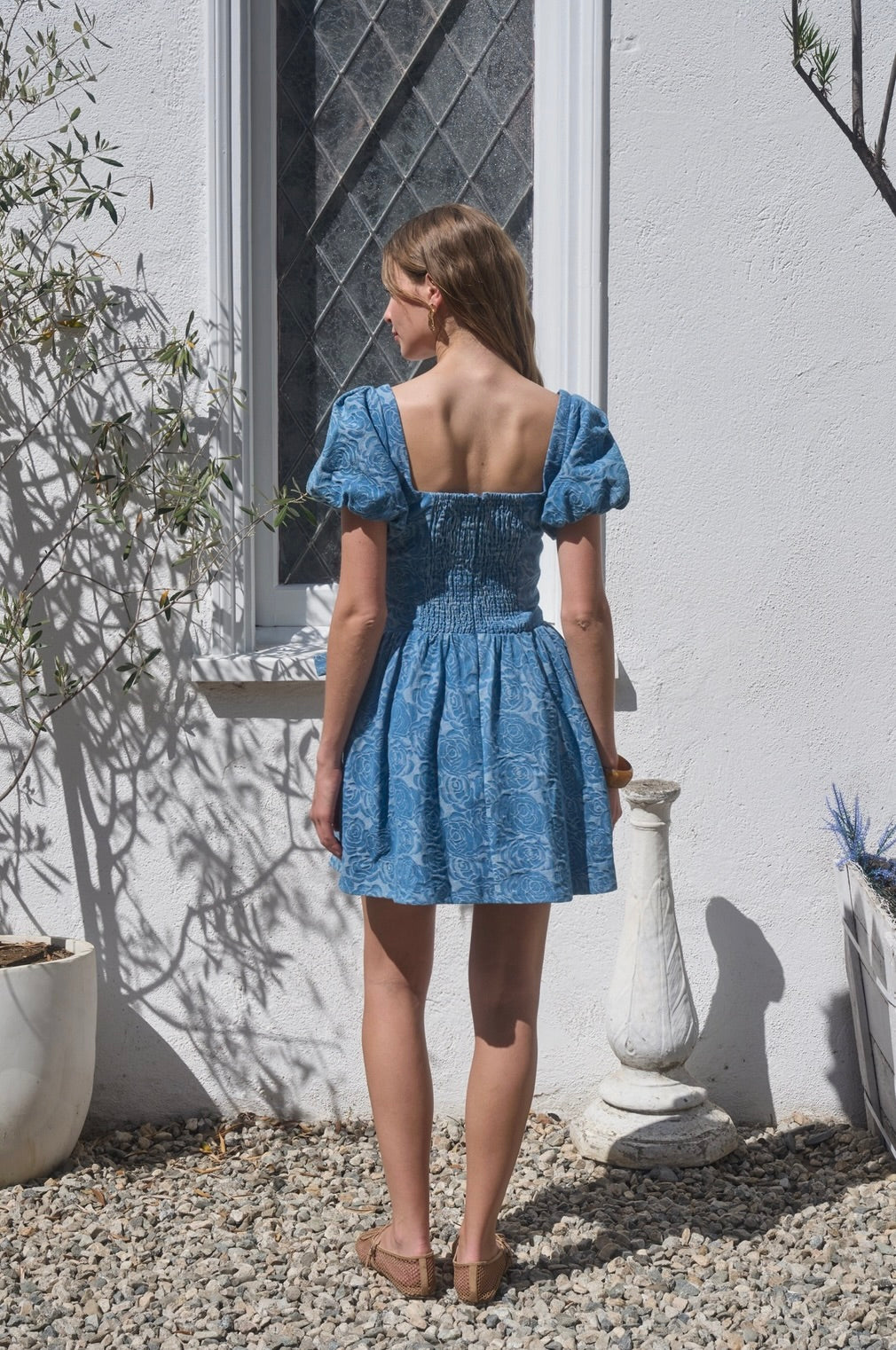 Woman wearing a blue dress standing outdoors against a white wall.