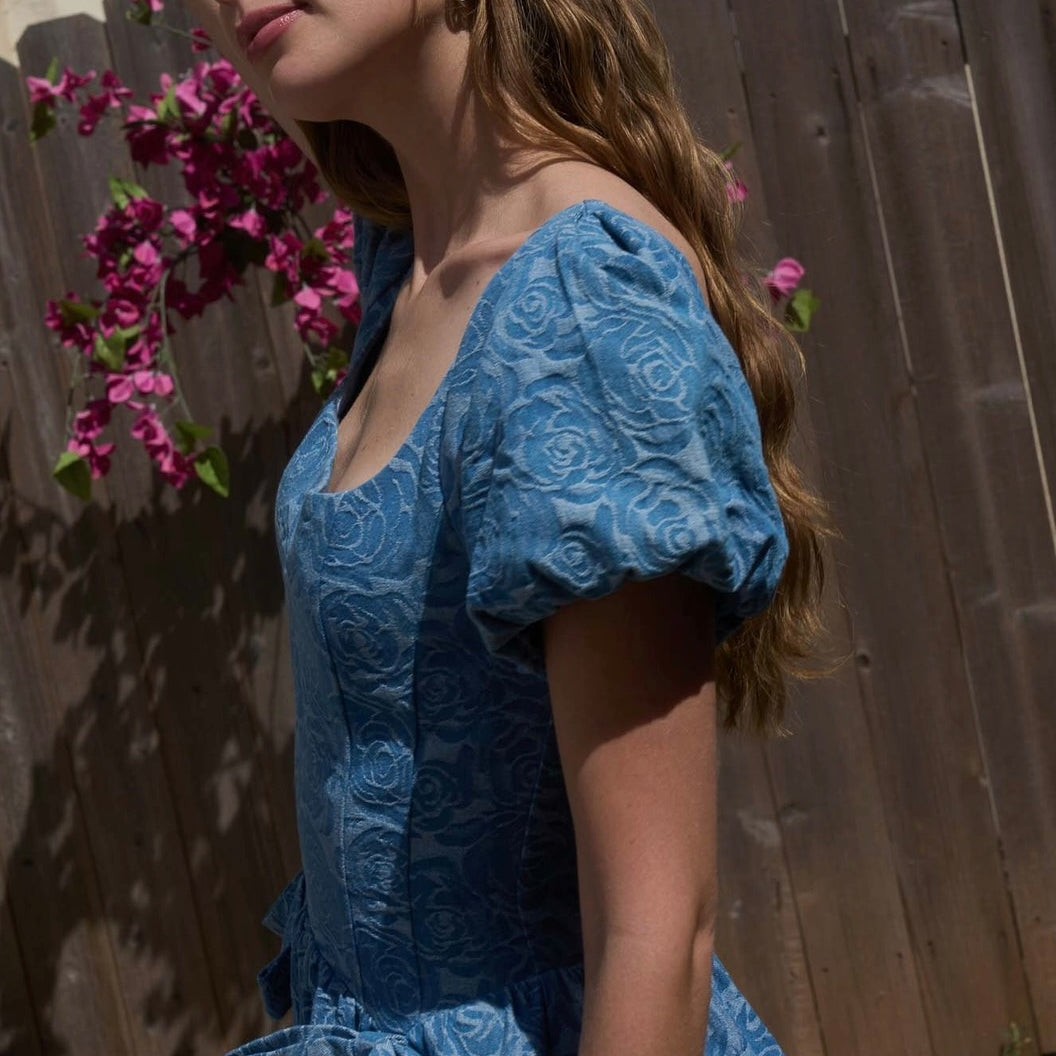 Woman in a blue lace dress standing outdoors with a wooden fence and flowers in the background