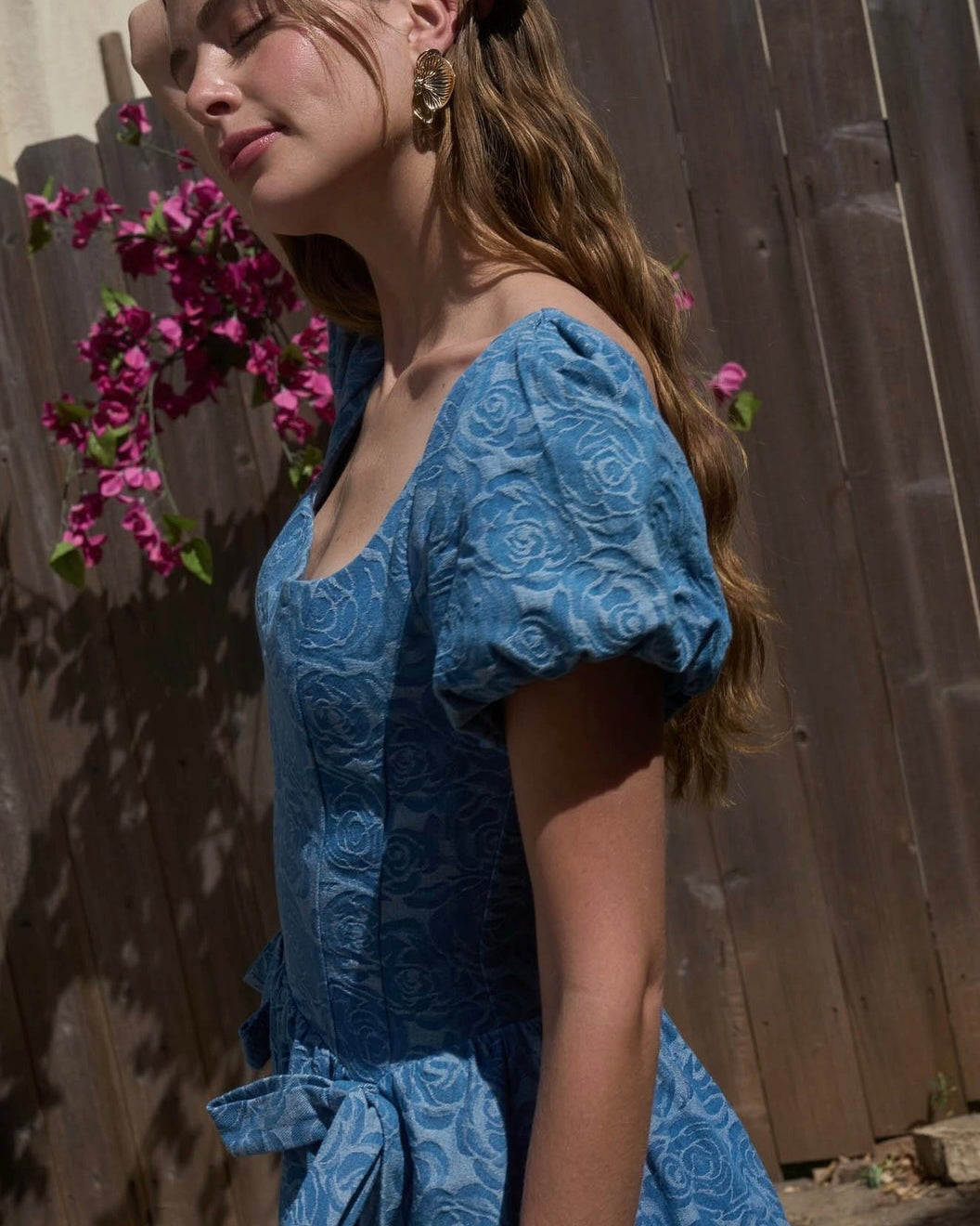Woman in a blue lace dress standing outdoors with a wooden fence and flowers in the background