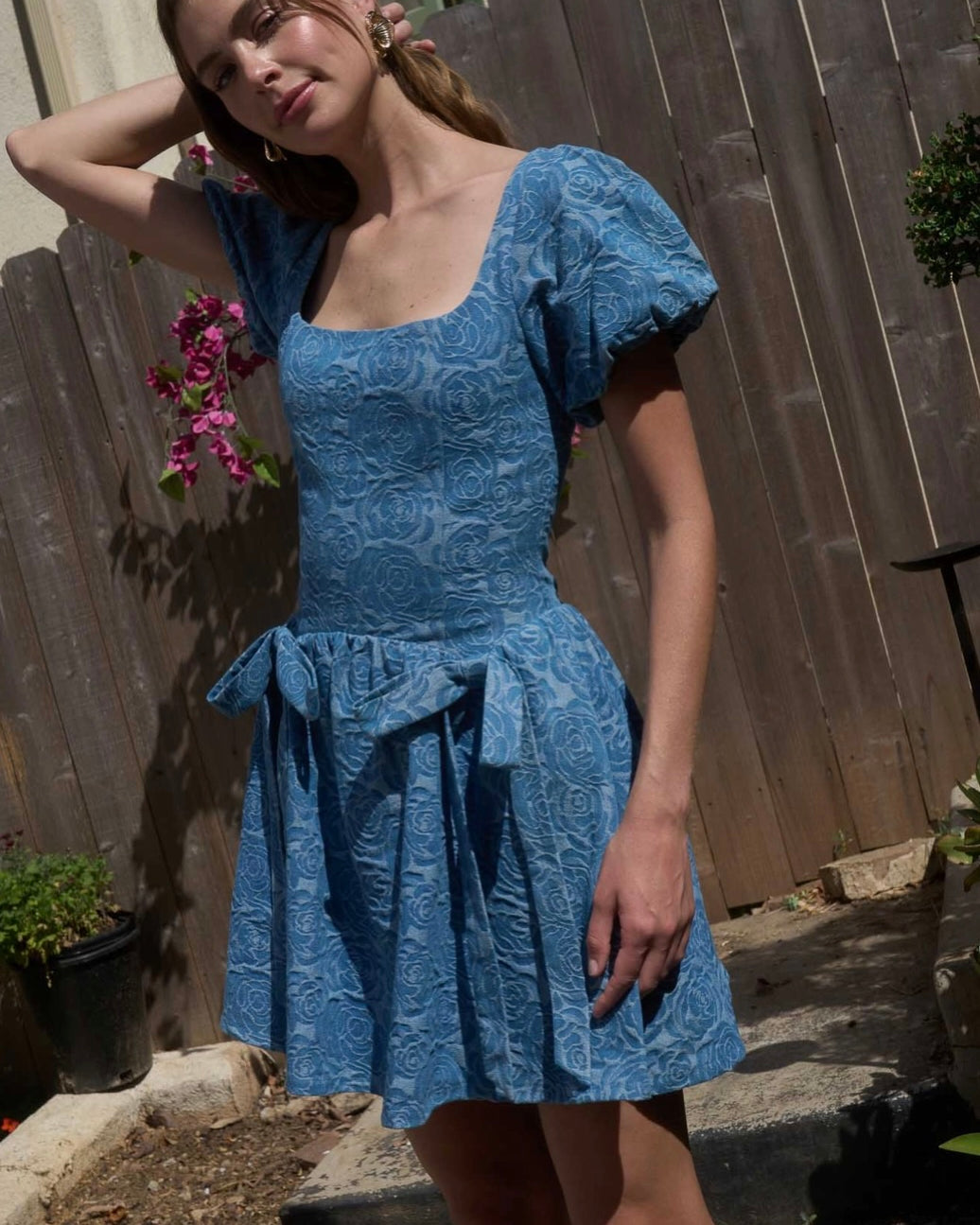 Woman wearing a blue dress standing outdoors with a wooden fence and plants in the background