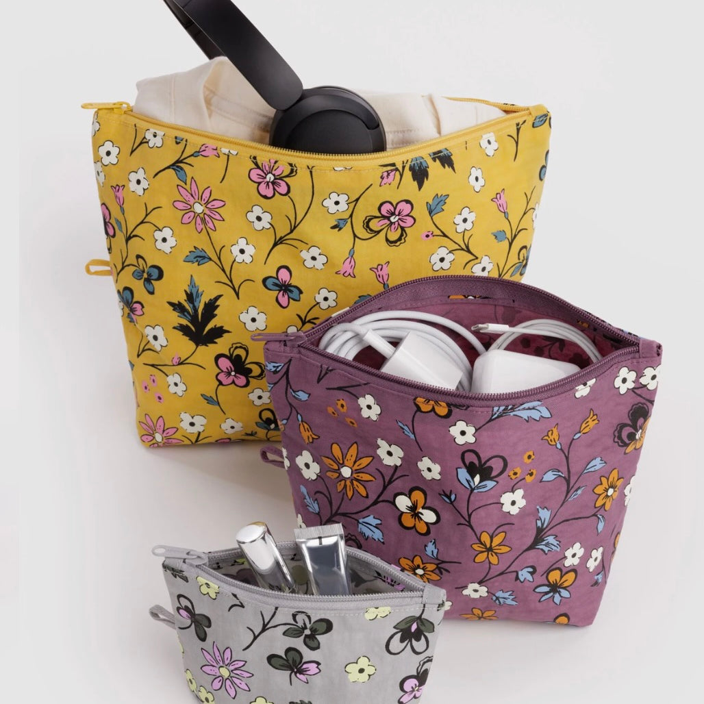 Three floral zippered cosmetic pouches in yellow, purple, and gray, shown with headphones, earbuds, and toiletries inside on a light gray background.