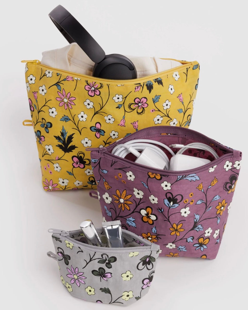 Three floral zippered cosmetic pouches in yellow, purple, and gray, shown with headphones, earbuds, and toiletries inside on a light gray background.