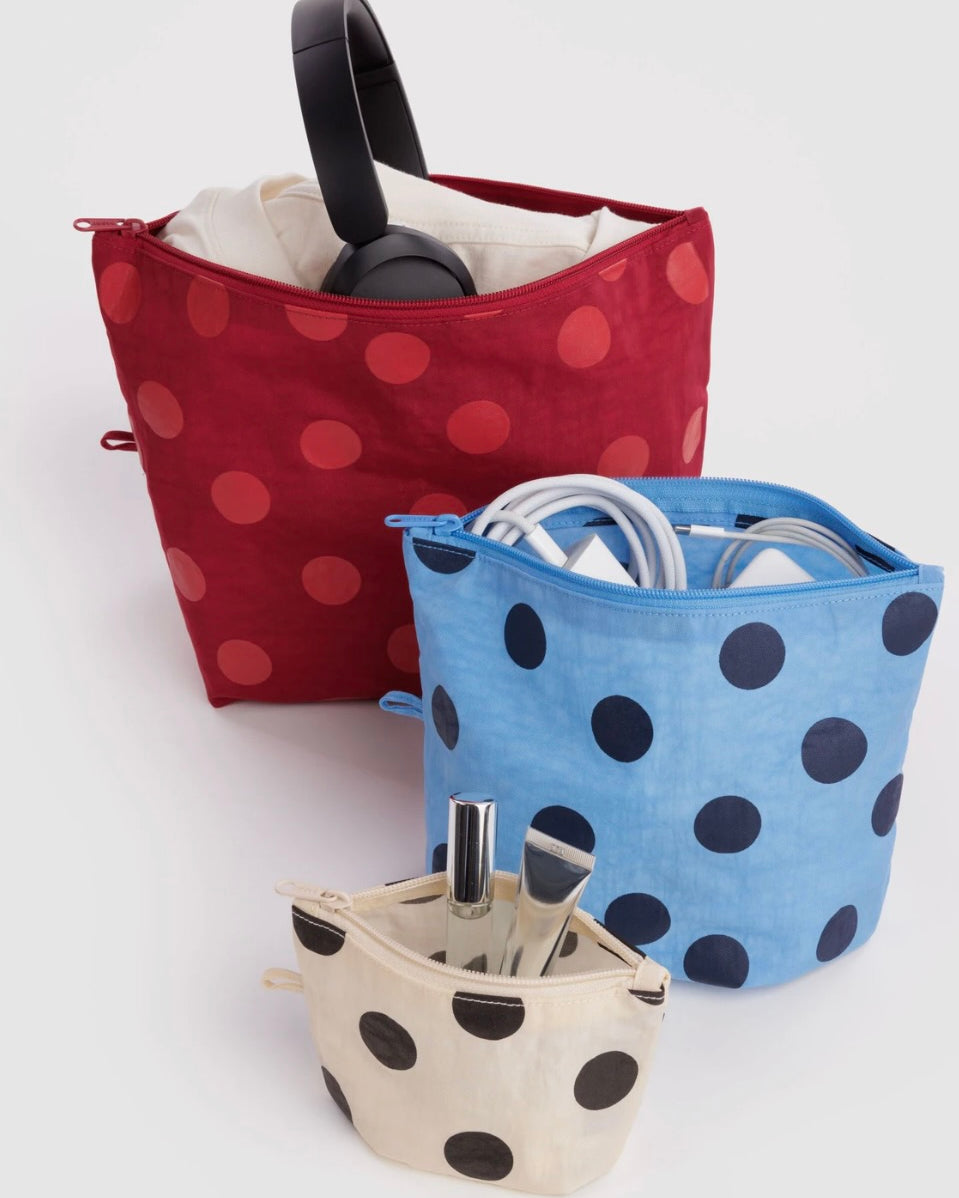 Three fabric zippered organizer pouches with polka dots, shown on a light gray studio background. The back pouch is dark red with red polka dots and contains black headphones; the middle pouch is light blue with dark blue polka dots and contains white cords; the front pouch is cream with black polka dots and contains a silver umbrella and pen.
