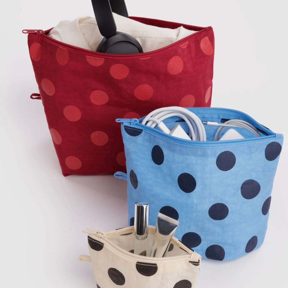 Three fabric zippered organizer pouches with polka dots, shown on a light gray studio background. The back pouch is dark red with red polka dots and contains black headphones; the middle pouch is light blue with dark blue polka dots and contains white cords; the front pouch is cream with black polka dots and contains a silver umbrella and pen.