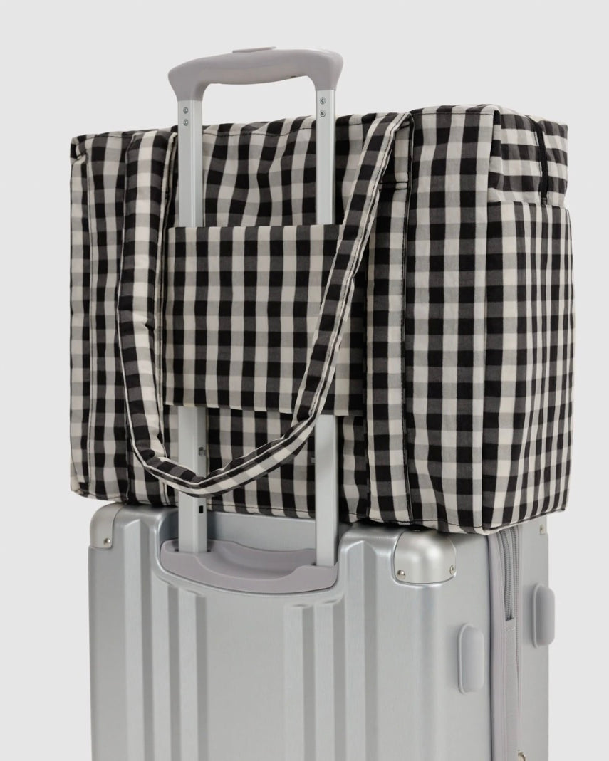 A black and white gingham checkered travel tote bag with a telescoping handle, shown resting on top of a silver hard-shell suitcase against a light gray background.