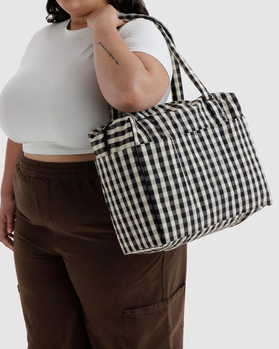 A person wearing a white crop top and brown pants carries a large black and white gingham check tote bag over their shoulder against a light gray background.