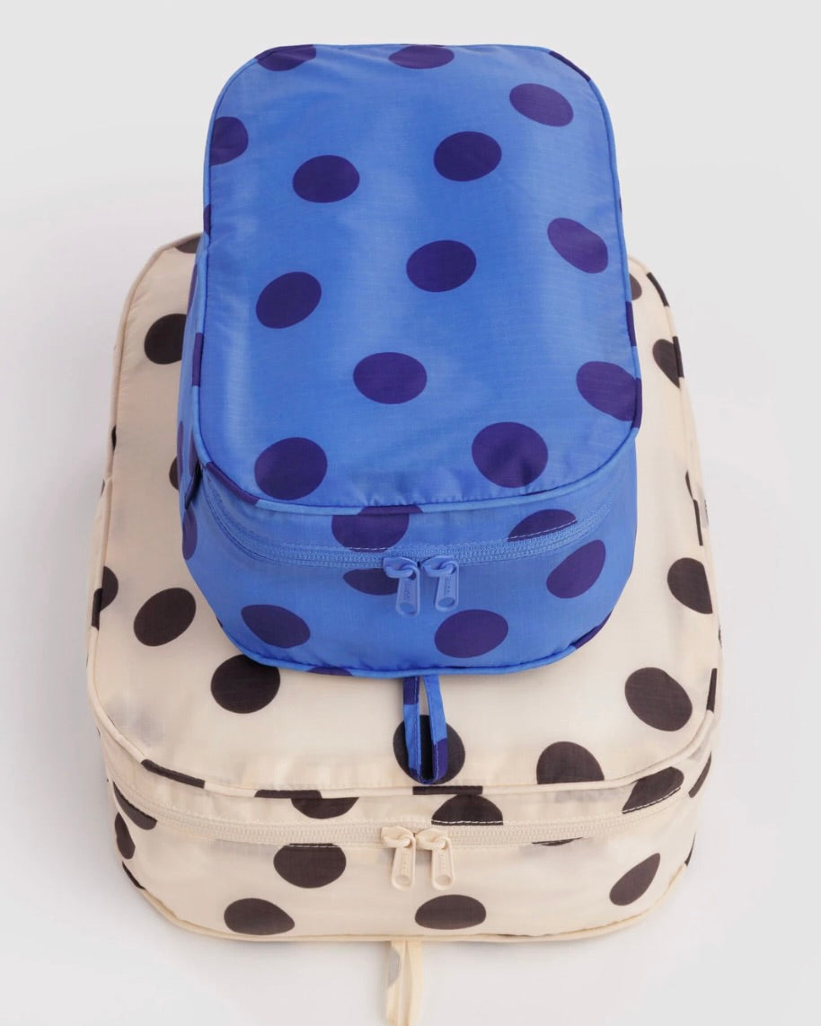 Two polka dot packing cubes stacked on a light gray background. The top cube is royal blue with dark purple polka dots, and the bottom cube is cream with large black polka dots.