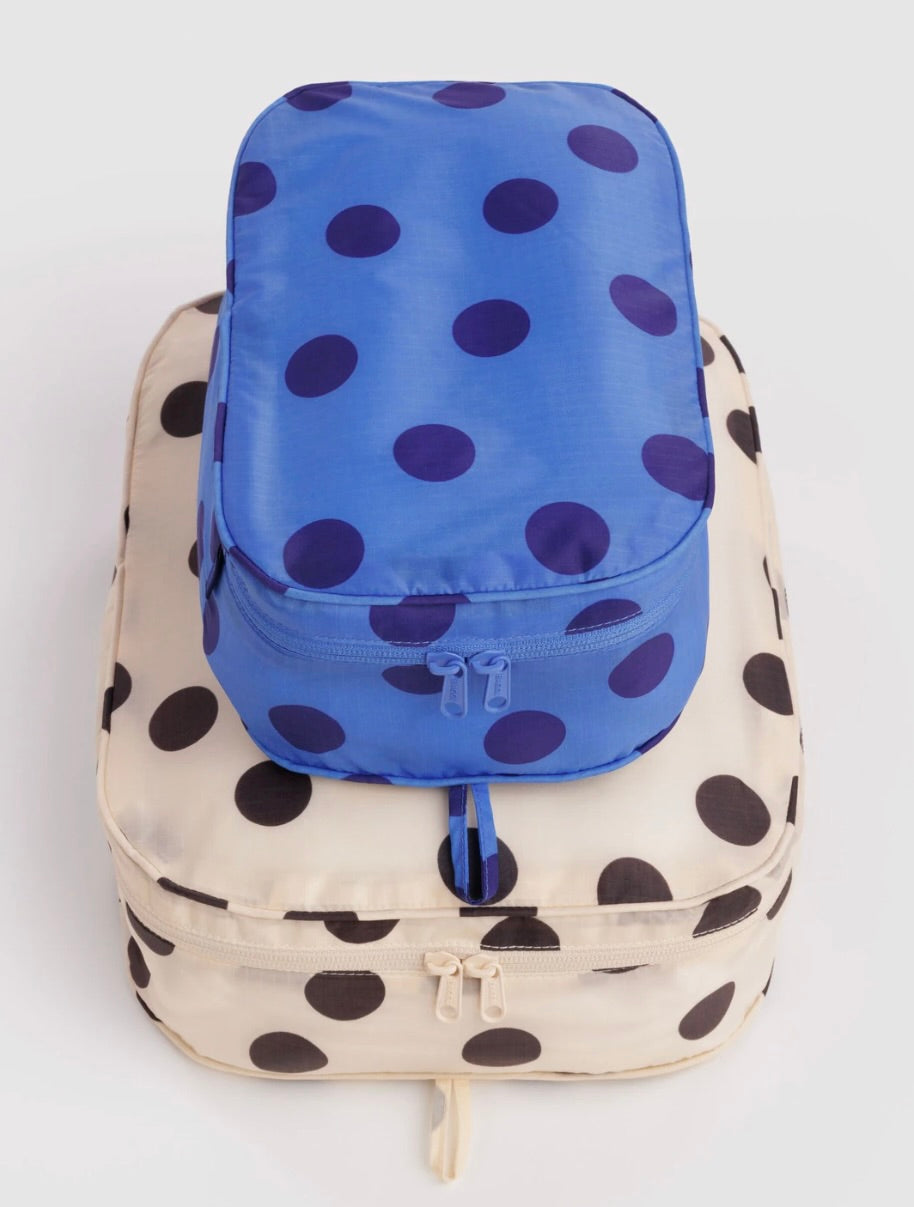Two polka dot packing cubes stacked on a light gray background. The top cube is royal blue with dark purple polka dots, and the bottom cube is cream with large black polka dots.