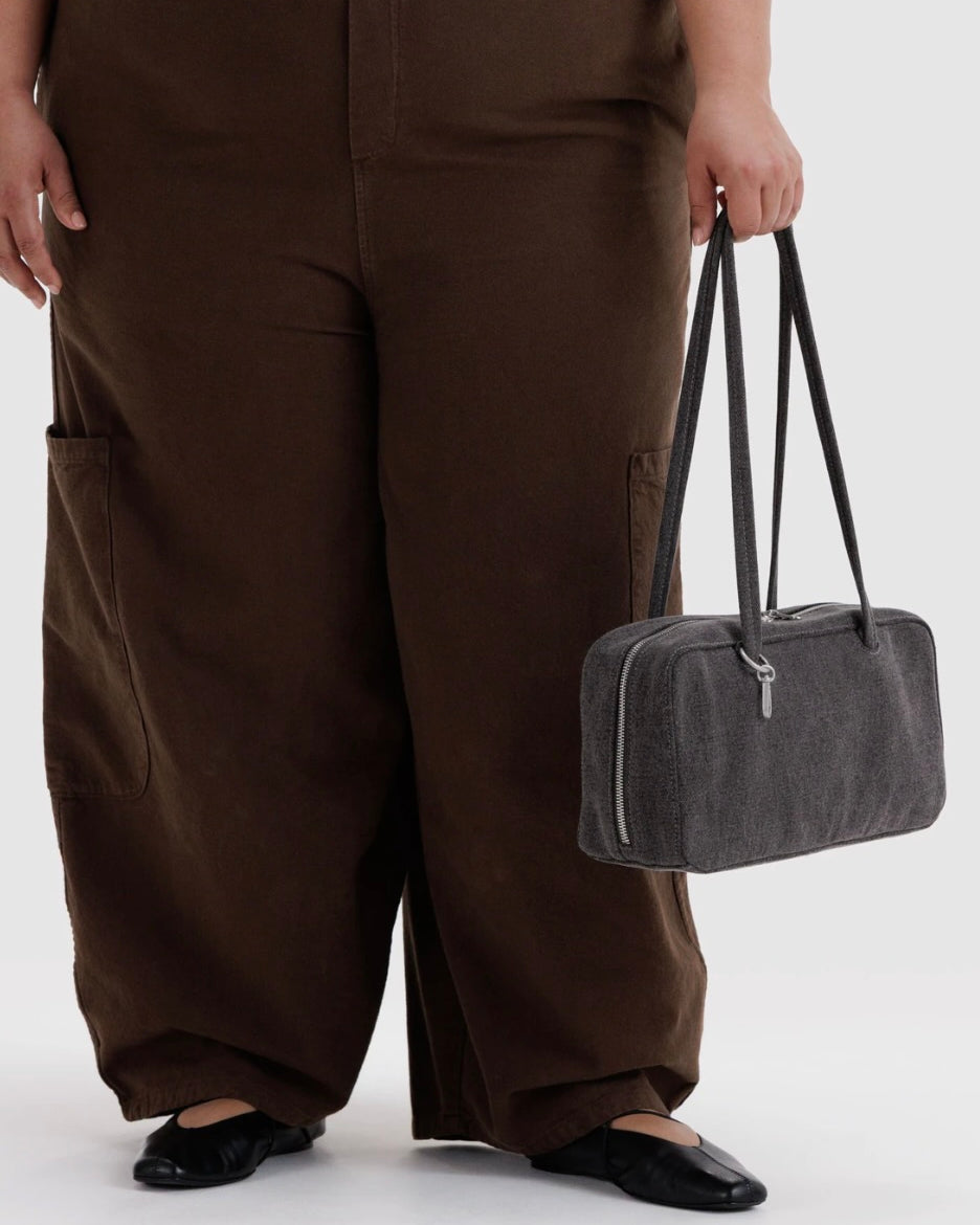 A person wearing loose brown cargo-style pants and black flats holds a dark gray rectangular shoulder bag with long handles against a light gray studio background.