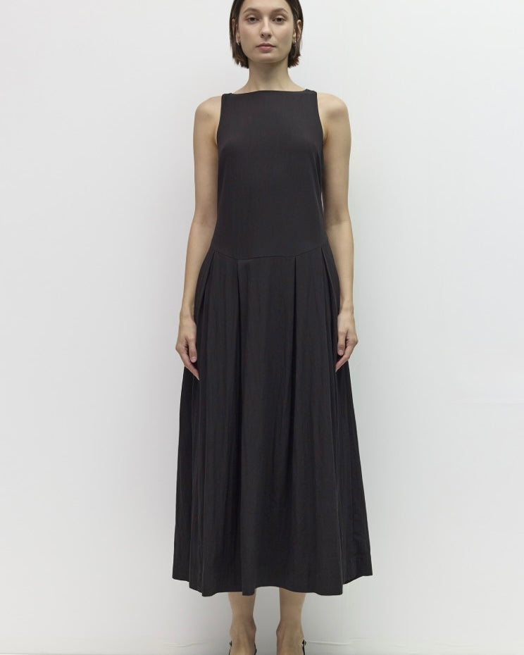 A woman stands against a plain white studio background wearing a sleeveless black midi dress with a structured bodice and flowing skirt, paired with black strappy heels.