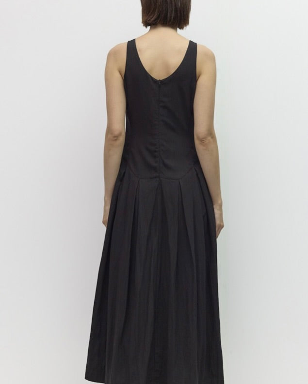 A woman viewed from the back wearing a sleeveless black midi dress with a V-neckline and gathered skirt, standing against a plain light gray wall.