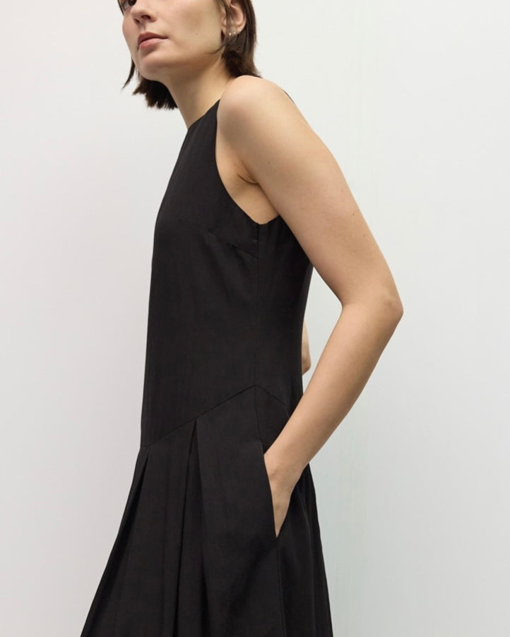 A woman wearing a sleeveless black midi dress with side pockets, photographed against a plain light gray studio background.