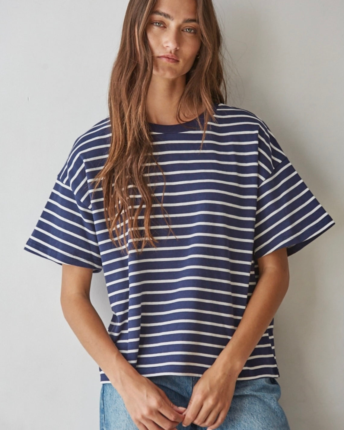 A woman wearing an oversized short-sleeve t-shirt with navy blue and white horizontal stripes, paired with light blue jeans, standing against a plain light gray wall.