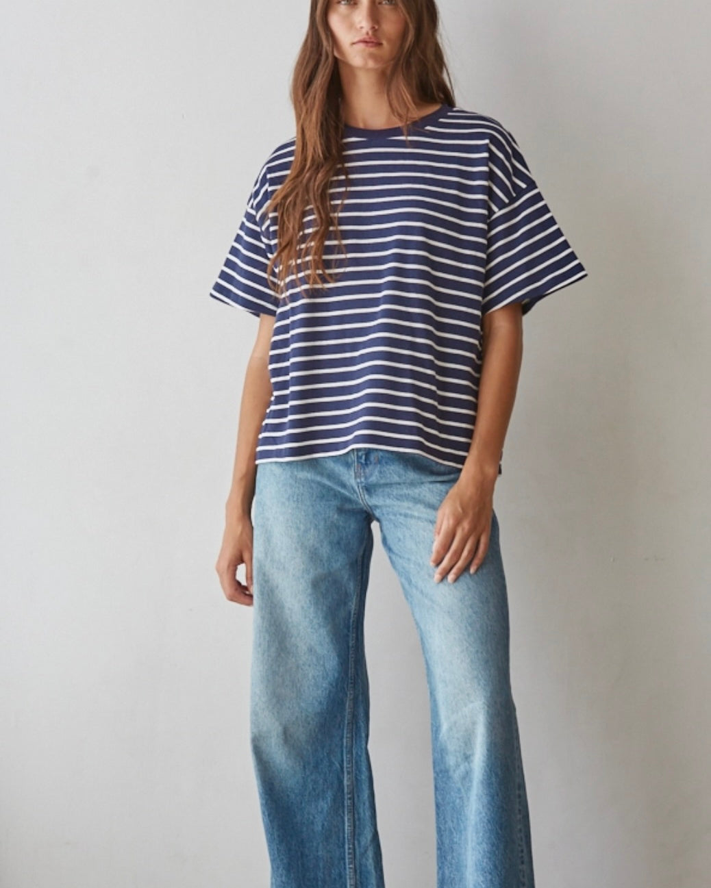 A woman modeling a navy blue and white horizontally striped short-sleeve t-shirt with light-wash blue wide-leg jeans, standing against a plain off-white wall.
