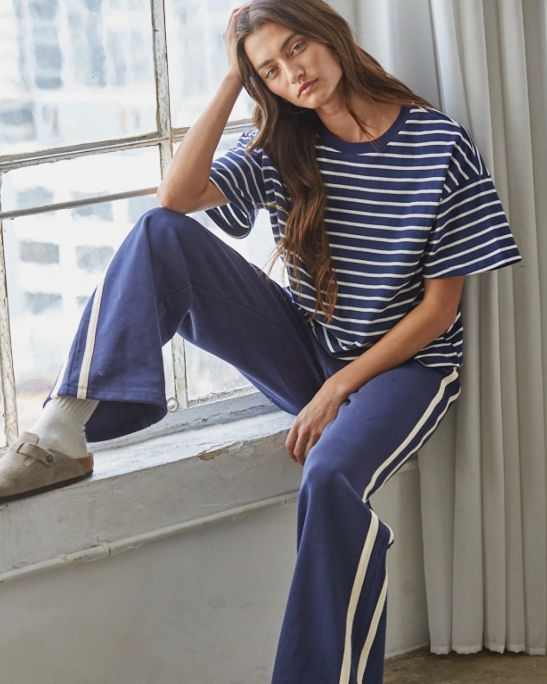 A woman sitting on a window sill wearing a navy blue and white striped short-sleeve t-shirt and matching navy blue wide-leg sweatpants with white side stripes.