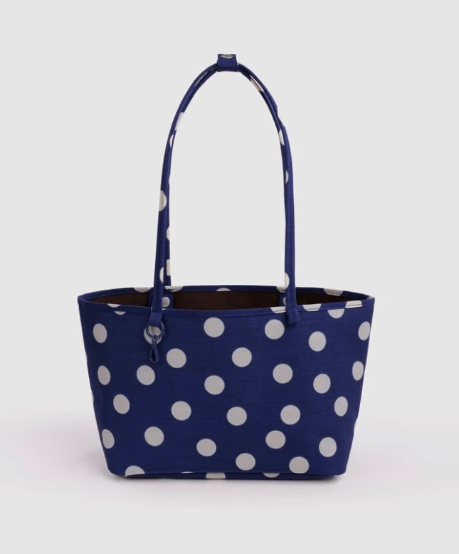 A navy blue canvas tote bag with a white polka dot pattern, featuring two shoulder straps and a small silver metal zipper pull on the left side, set against a plain light gray background.