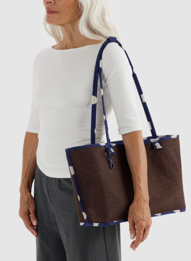 A woman wearing a white top and gray pants carries a large brown tote bag with navy and white patterned straps and trim against a light gray studio background.