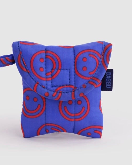 A small quilted pouch in bright blue with a red smiley face pattern, featuring a top flap and a small black brand tag that reads BAGGU.