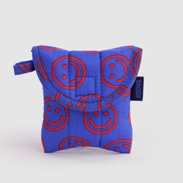 A small quilted pouch in bright blue with a red smiley face pattern, featuring a top flap and a small black brand tag that reads BAGGU.