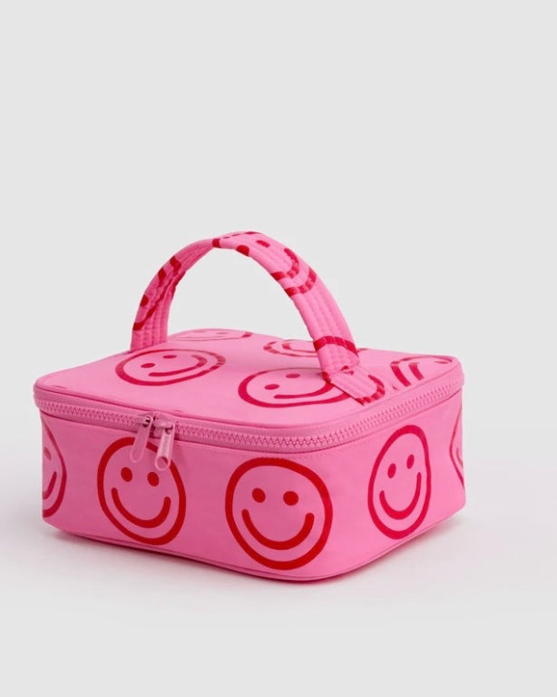 A pink zippered lunch bag with a top handle, featuring an all-over red smiley face pattern, photographed on a light gray background.