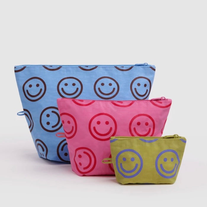 Three fabric zip pouches in blue, pink, and olive green, each printed with a repeating smiley face pattern in contrasting colors, arranged on a light gray background.