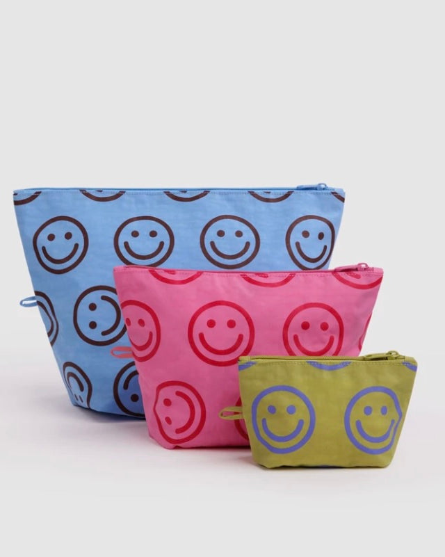 Three fabric zip pouches in blue, pink, and olive green, each printed with a repeating smiley face pattern in contrasting colors, arranged on a light gray background.