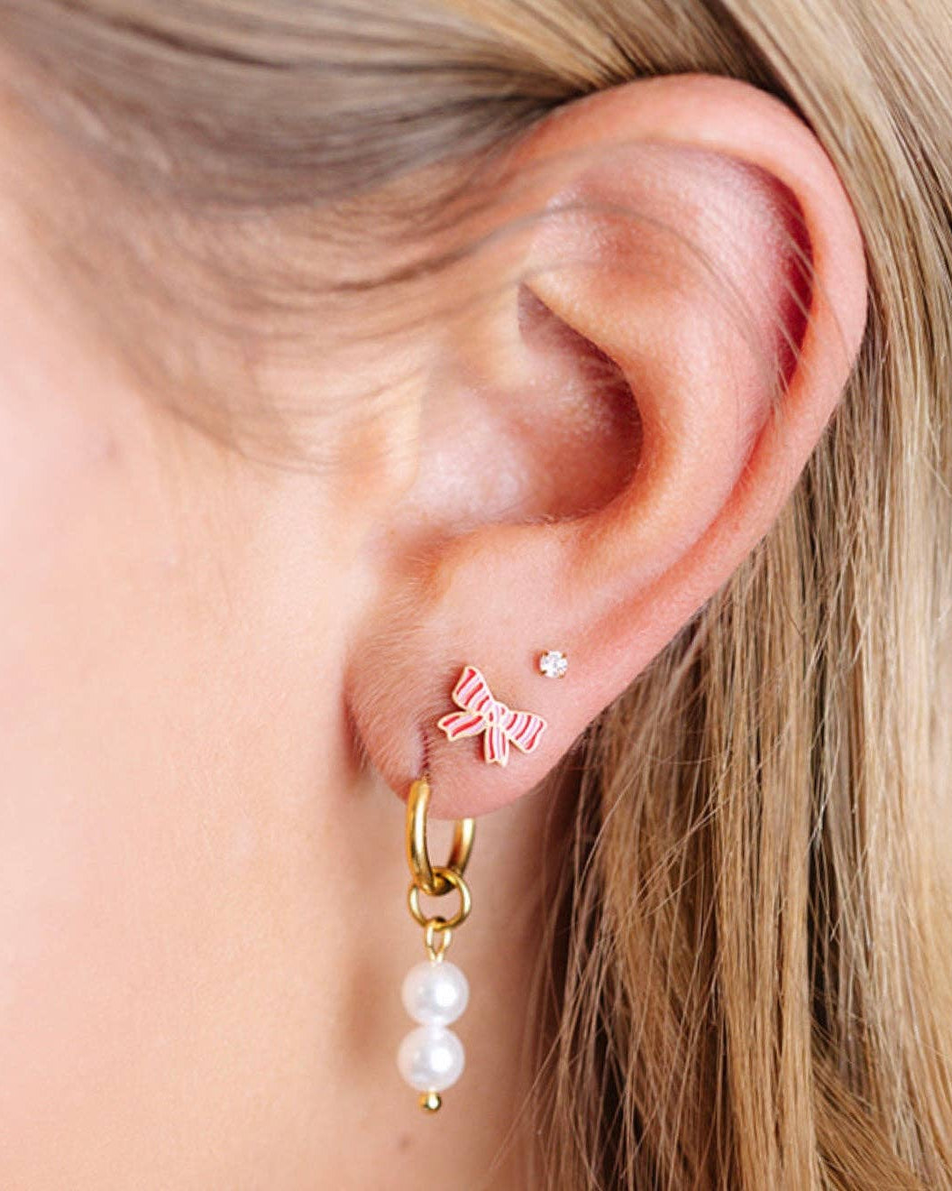 Close-up of an ear wearing a gold hoop earring with pearl drop and a small bow-shaped earring.