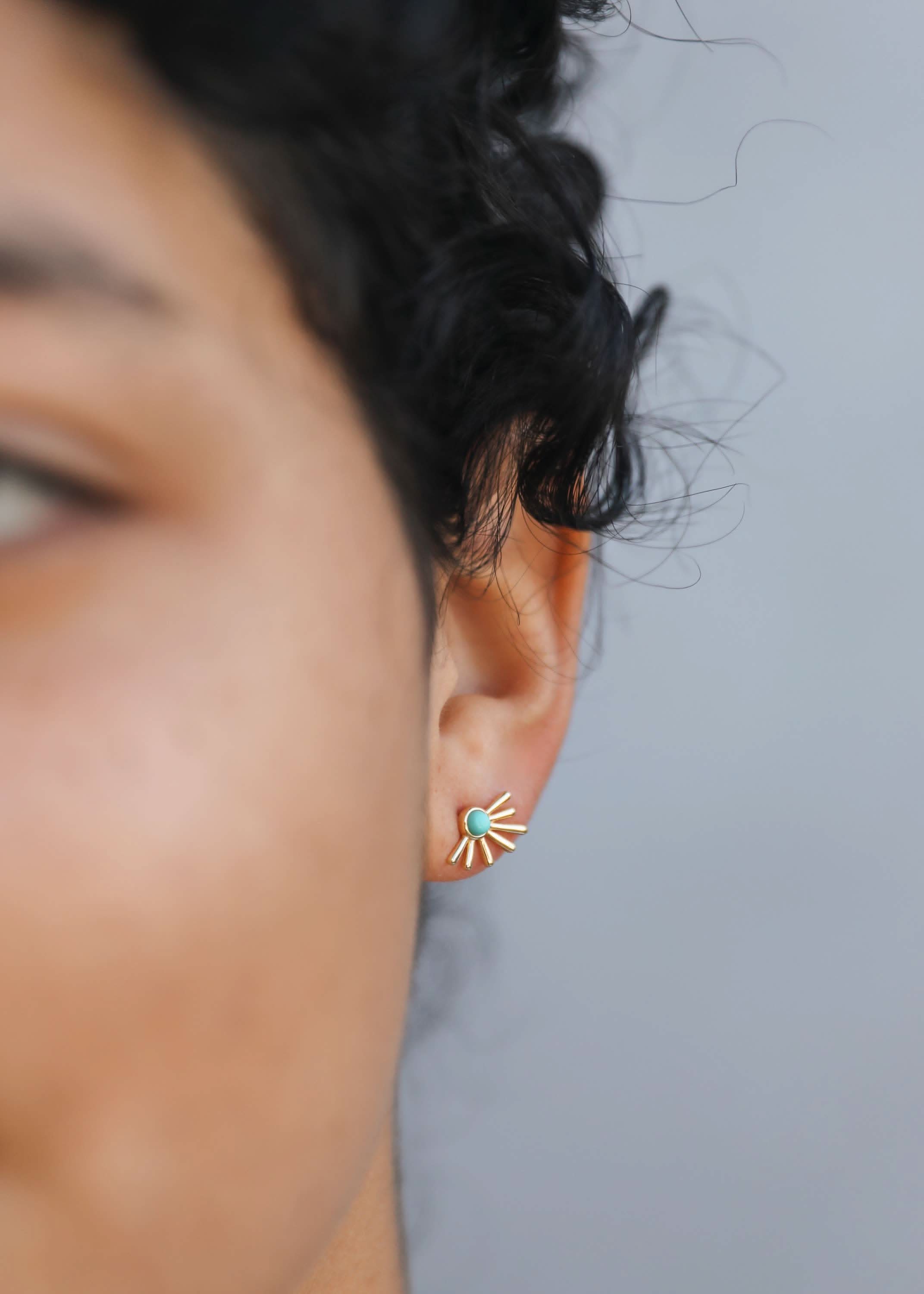 Close-up of an ear wearing a turquoise sun-shaped earring against a neutral background
