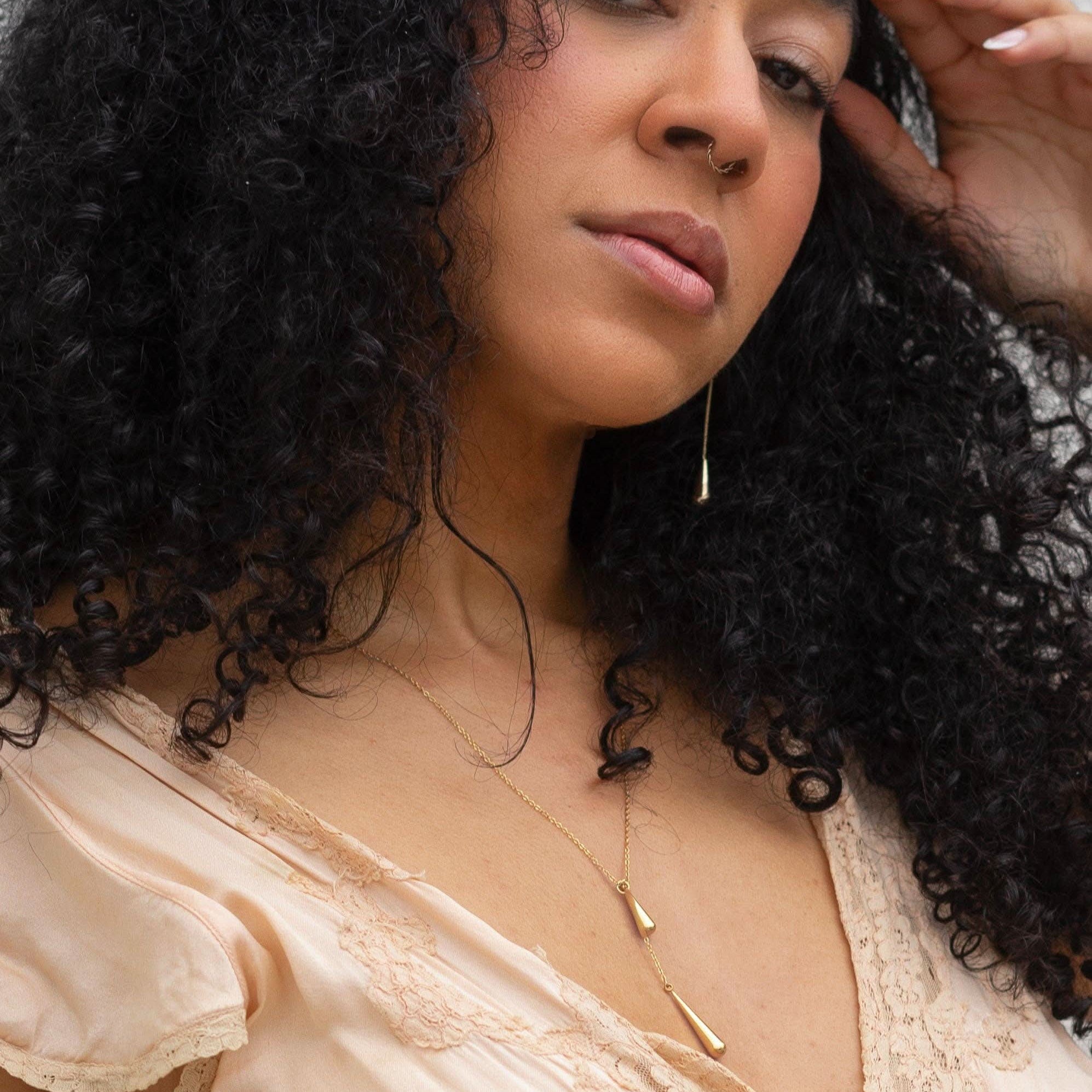 Woman with curly hair wearing a beige top and gold necklace against a neutral background