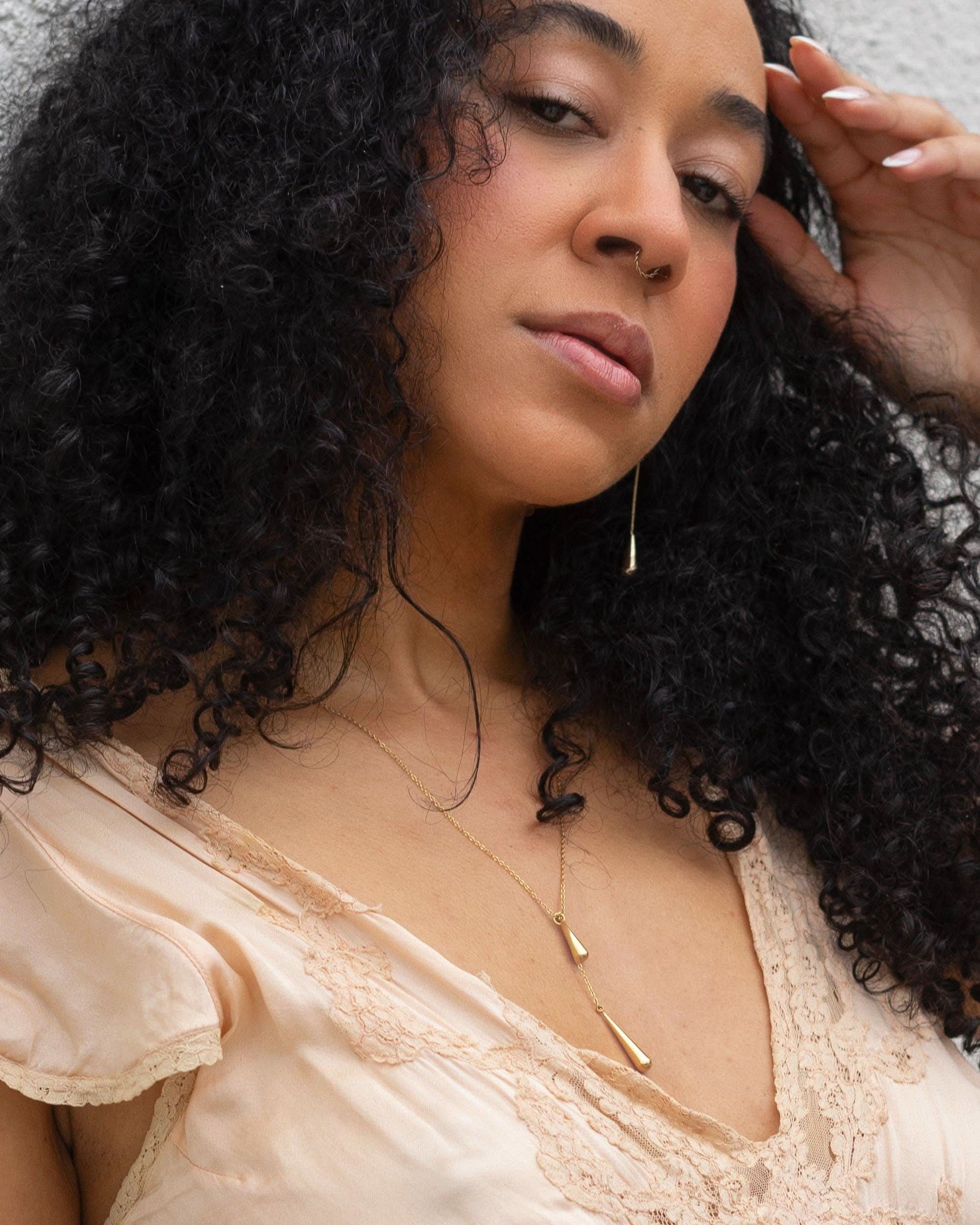 Woman with curly hair wearing a beige top and gold necklace against a neutral background