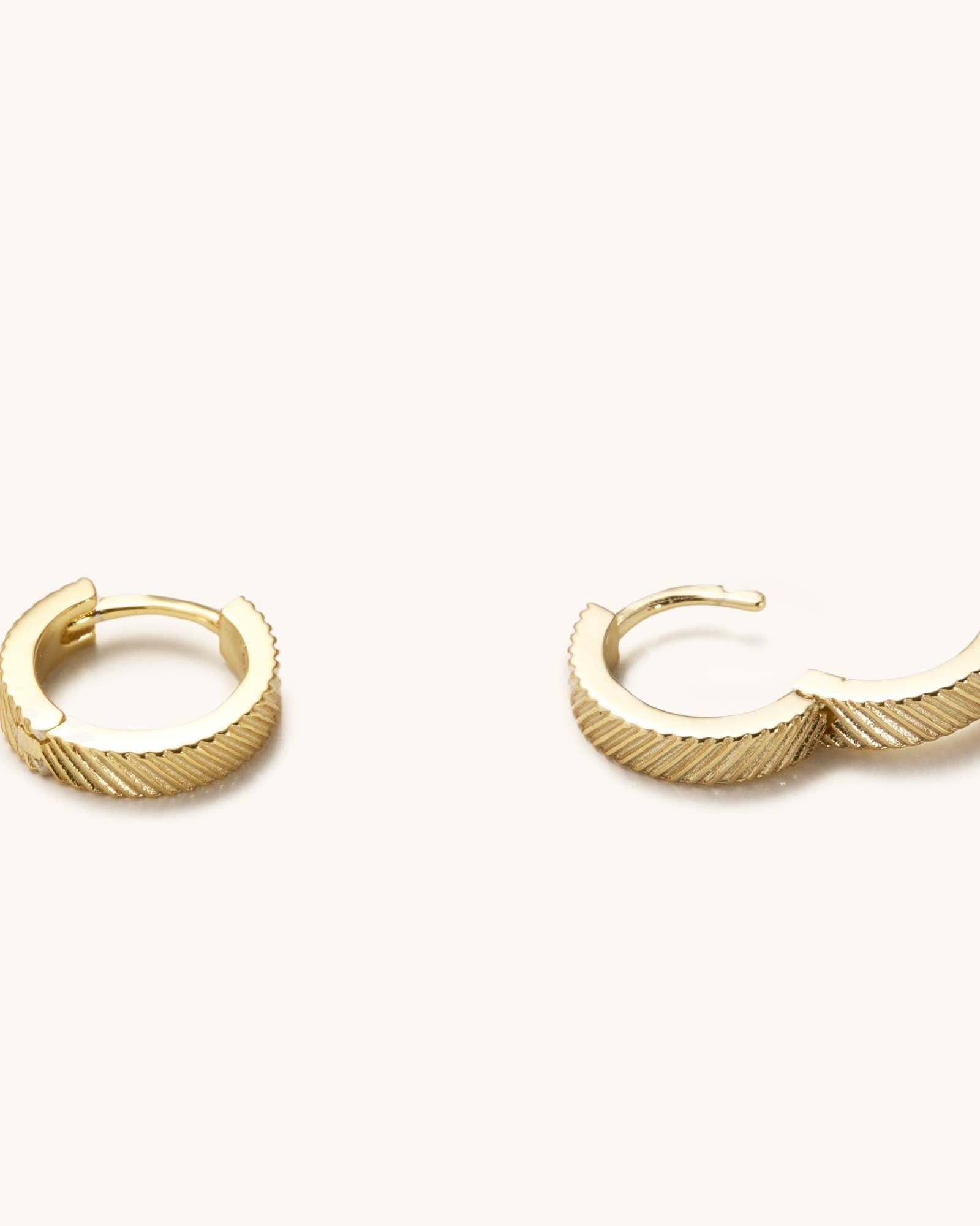 Pair of textured gold huggie loop hoop earrings laying on a white background