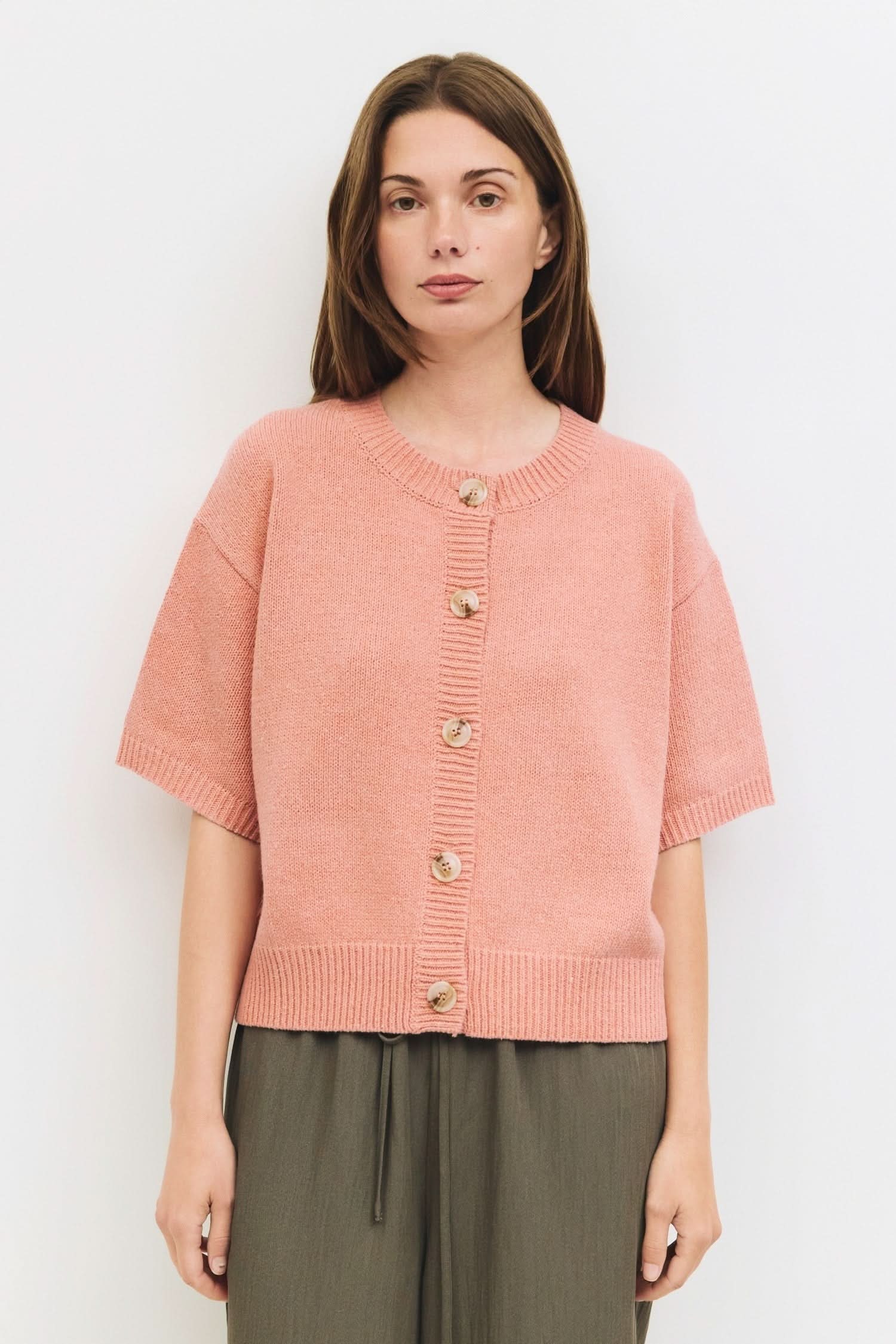 Woman wearing a salmon pink shirt sleeved button down cardigan standing against a white wall