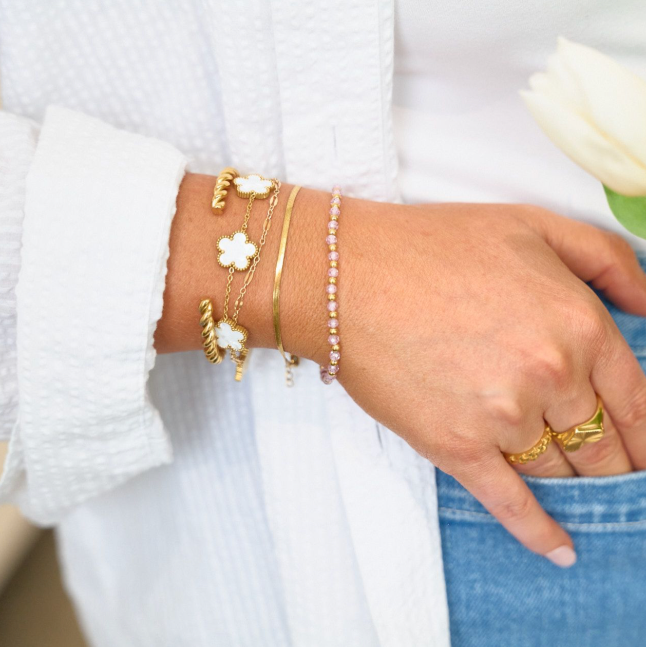 Hand wearing gold bracelets and a ring, holding a white tulip against