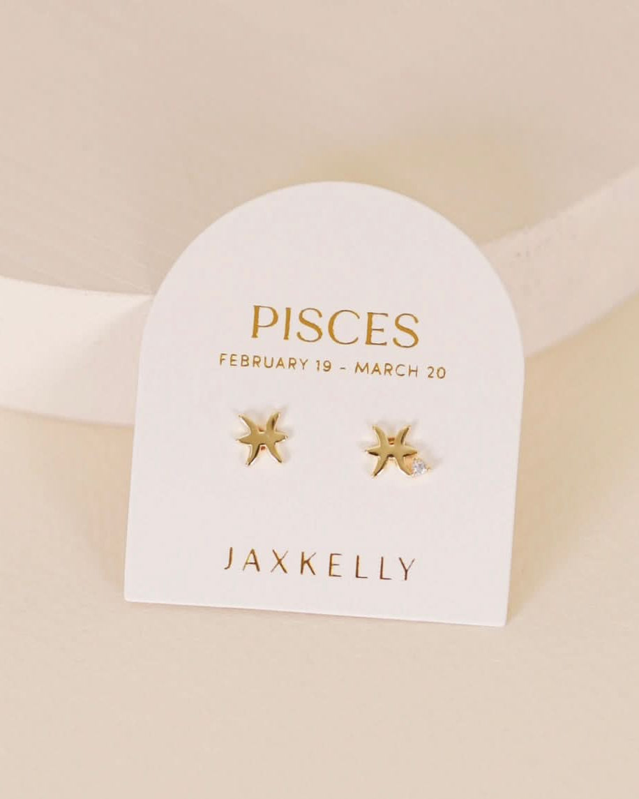 Gold zodiac charm studs in the shape of Pisces astrological sign displayed on earring card