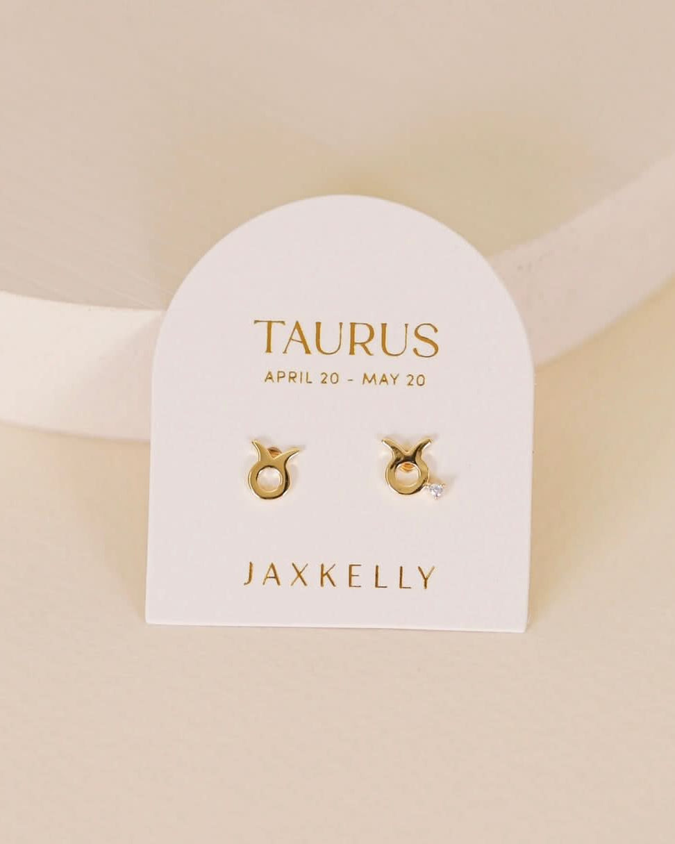 Gold zodiac charm studs in the shape of Taurus astrological sign displayed on earring card