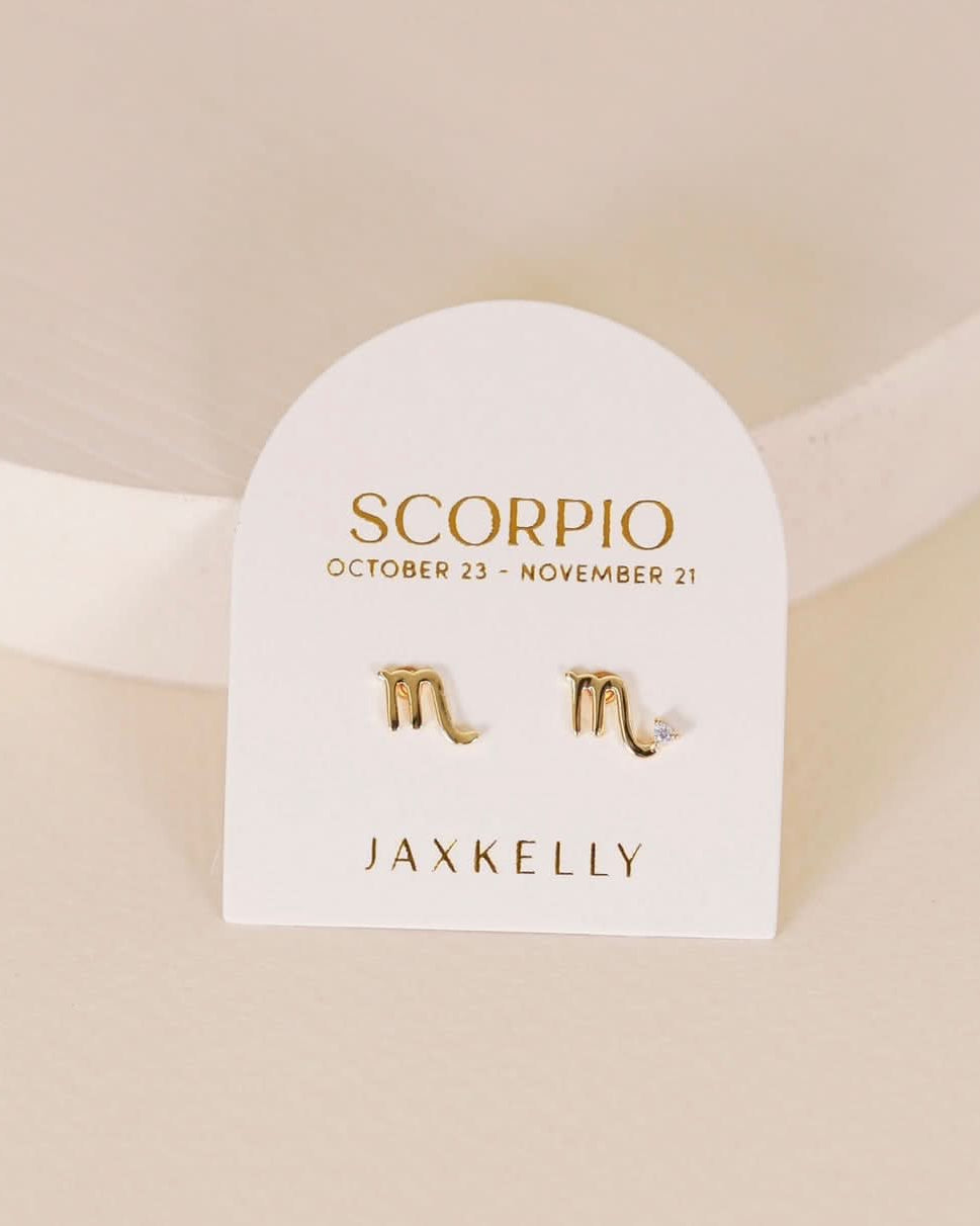 Gold zodiac charm studs in the shape of Scorpio astrological sign displayed on earring card