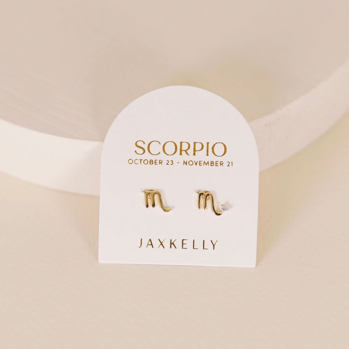 Gold zodiac charm studs in the shape of Scorpio astrological sign displayed on earring card